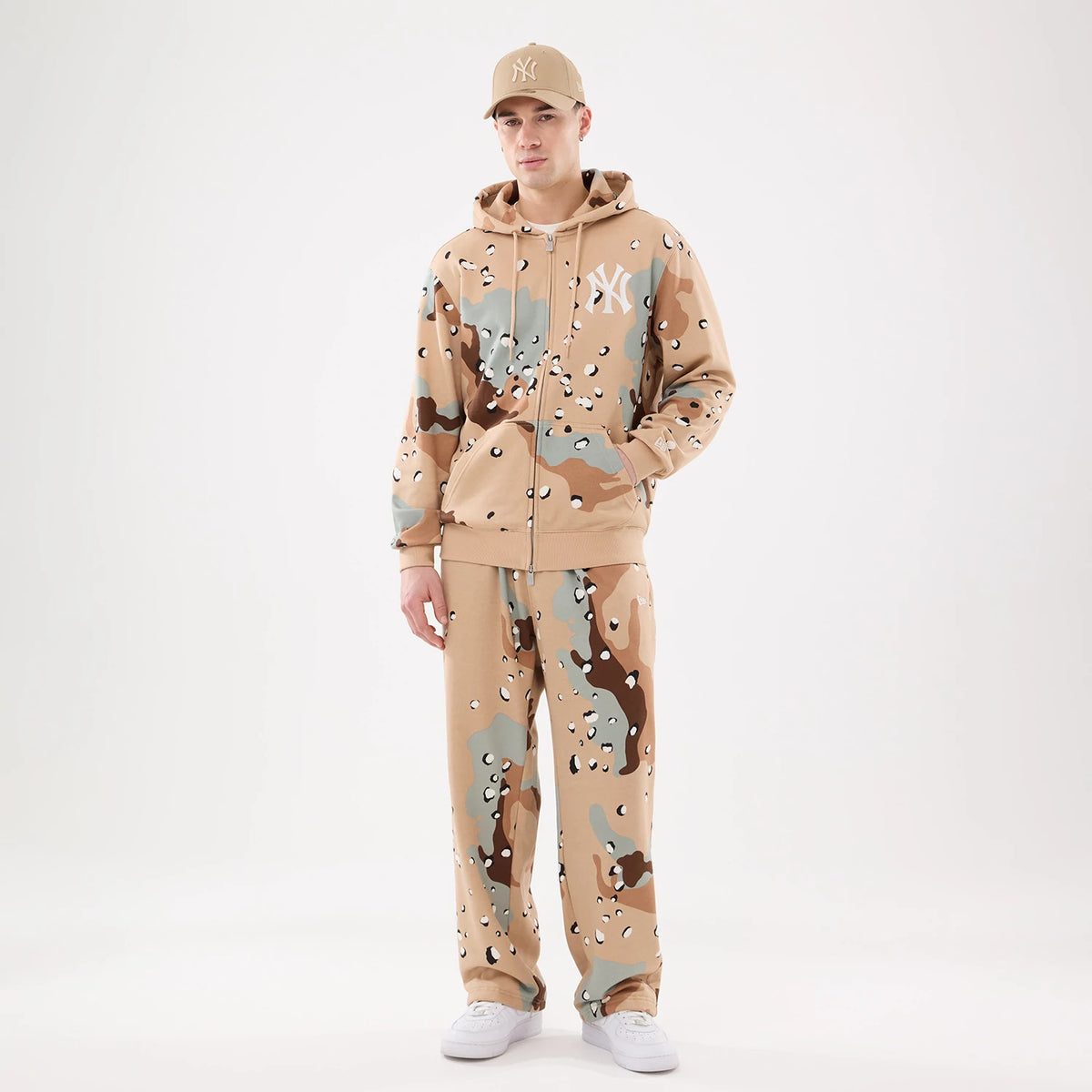 New Era Desert Camo Joggers, on-model with a matching hoodie in a tan, brown, and grey camouflage pattern, showing a relaxed fit and elasticated waistband, paired with white trainers and a camel cap.