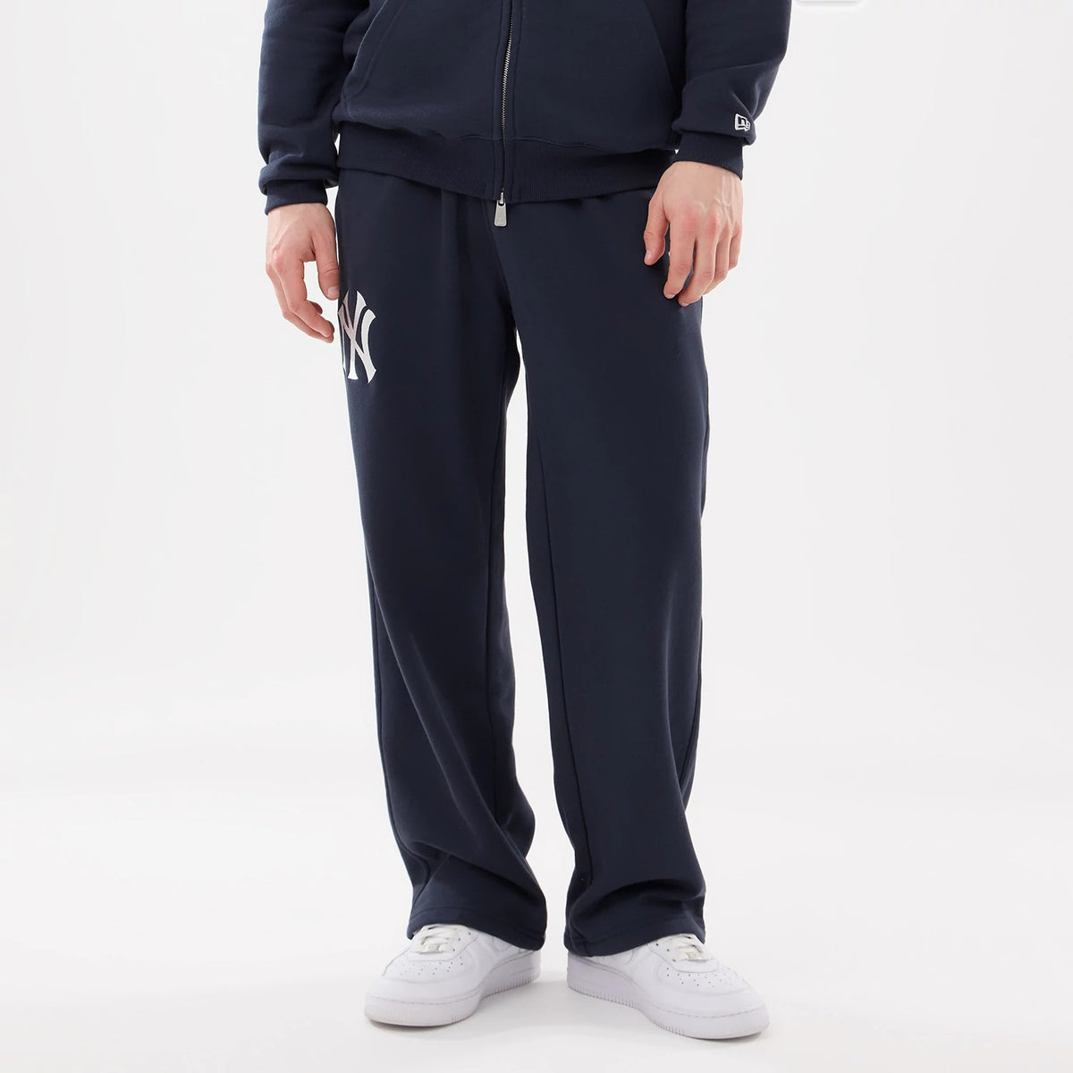 New York Yankees MLB Comfort Blue Joggers, front view on a model showing the white Yankees logo embroidered on the left leg, with an elastic waistband and straight-leg fit, paired with white trainers and a matching blue zip-up top.