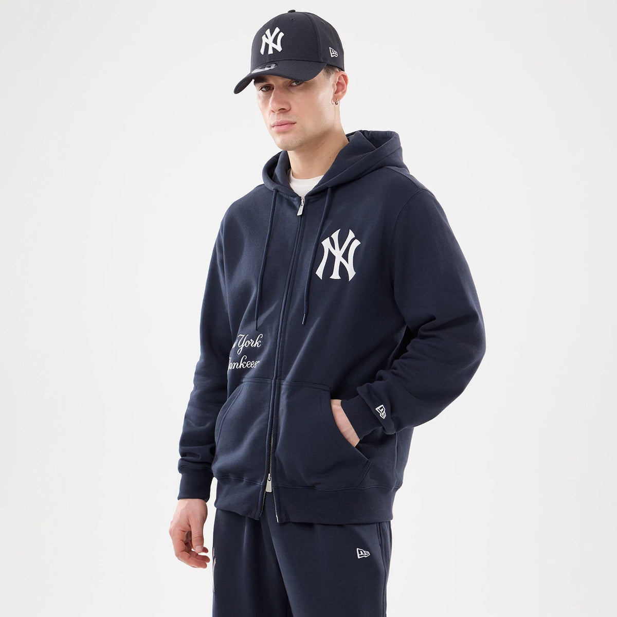 New York Yankees MLB Comfort Navy 2 Way Full Zip Hoodie, on-model view showing the navy hoodie with a white embroidered Yankees logo on the chest, full zip closure and pockets, paired with a matching navy cap featuring the same logo.