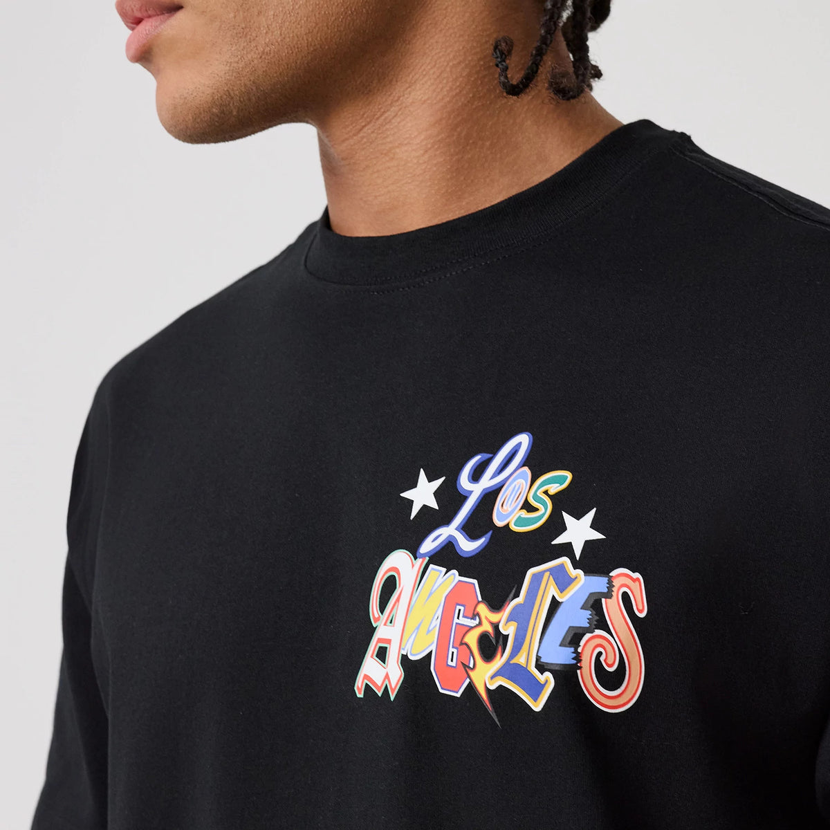 New Era LA Dodgers MLB Word Art Black T-Shirt, on-model view showing a colourful printed design with Los Angeles text and star accents on the upper chest of the solid black t-shirt.