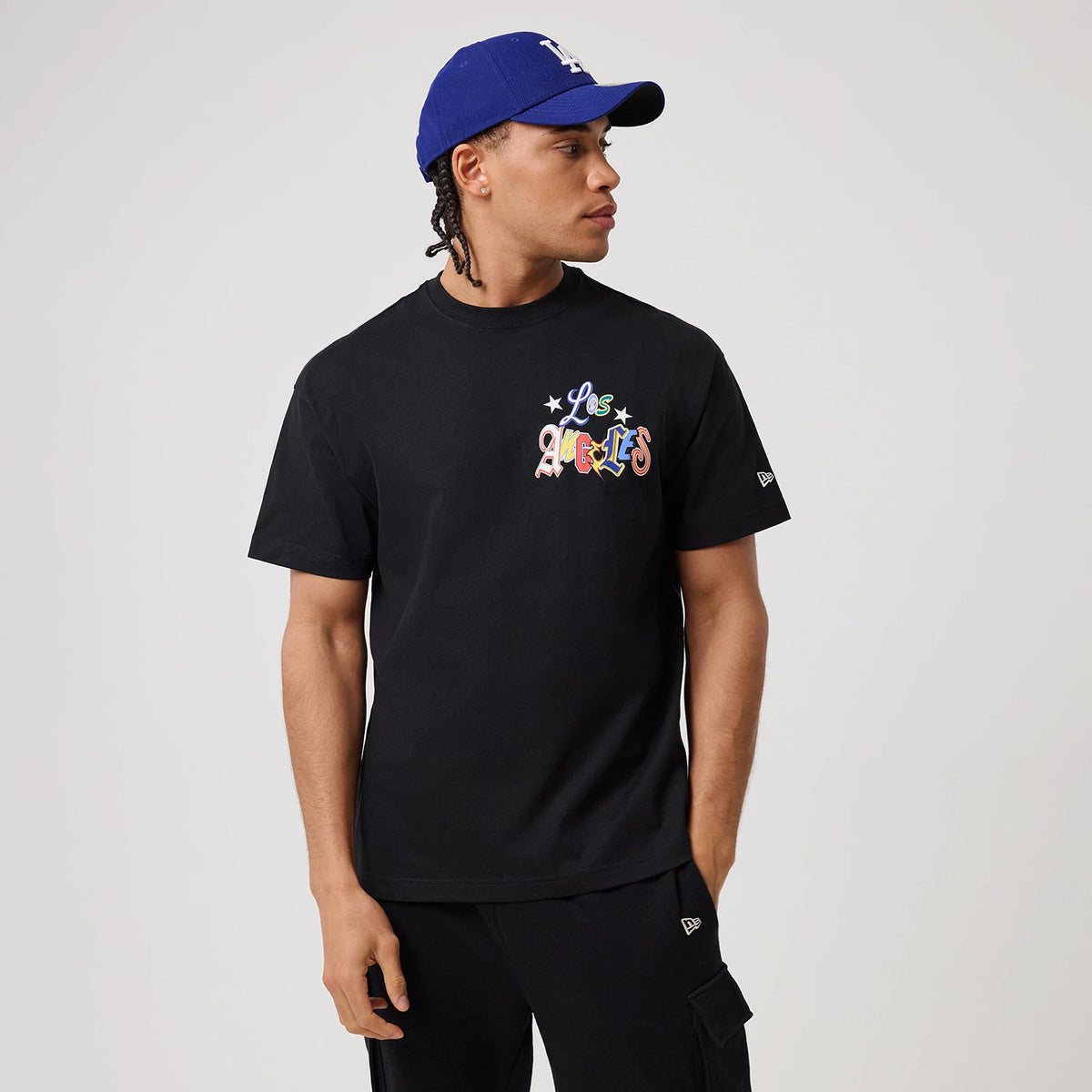 LA Dodgers MLB Word Art Black T-Shirt, on-model view showing the colourful text design on the chest with stars, alongside a small raised New Era logo on the sleeve, paired with a purple cap with a raised LA logo.
