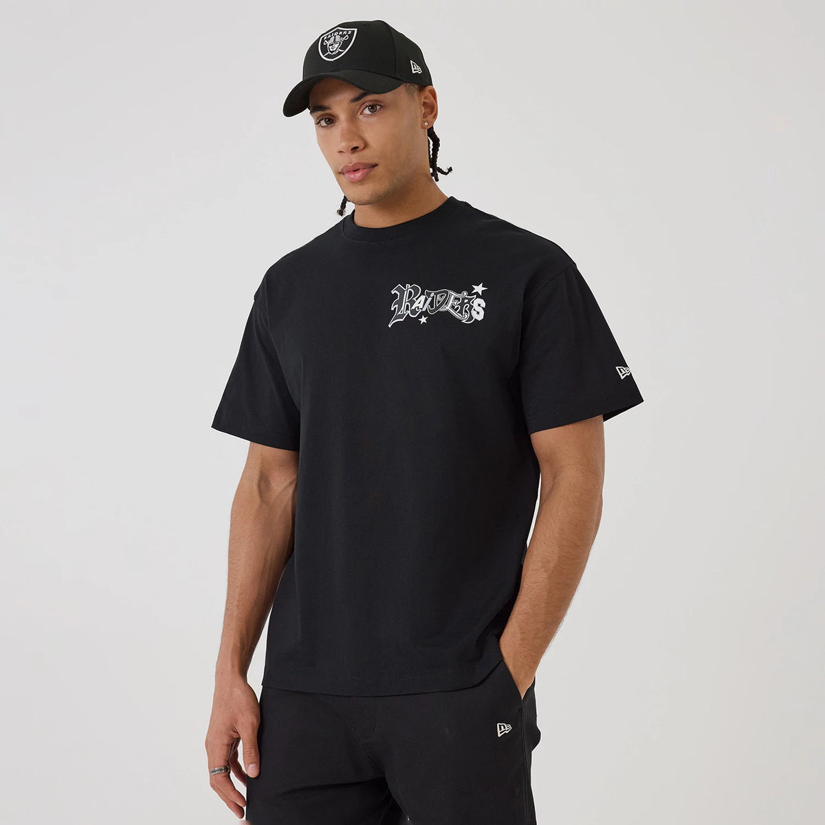 Las Vegas Raiders NFL Word Art Black T-Shirt, on-model view showing the black shirt with a raised "Raiders" logo on the chest, a New Era logo on the left sleeve, and a black cap featuring a raised Raiders logo on the front.