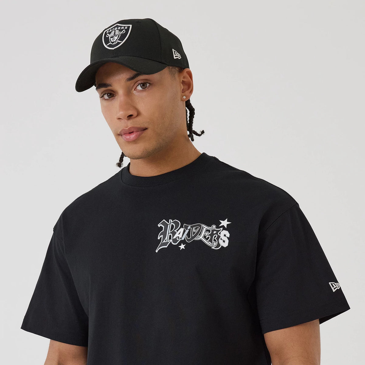 Las Vegas Raiders NFL Word Art Black T-Shirt, front view showing the black shirt with "Raiders" text and star graphics on the chest, a small New Era logo on the left sleeve, worn with a black cap featuring an embroidered Raiders logo on the front.