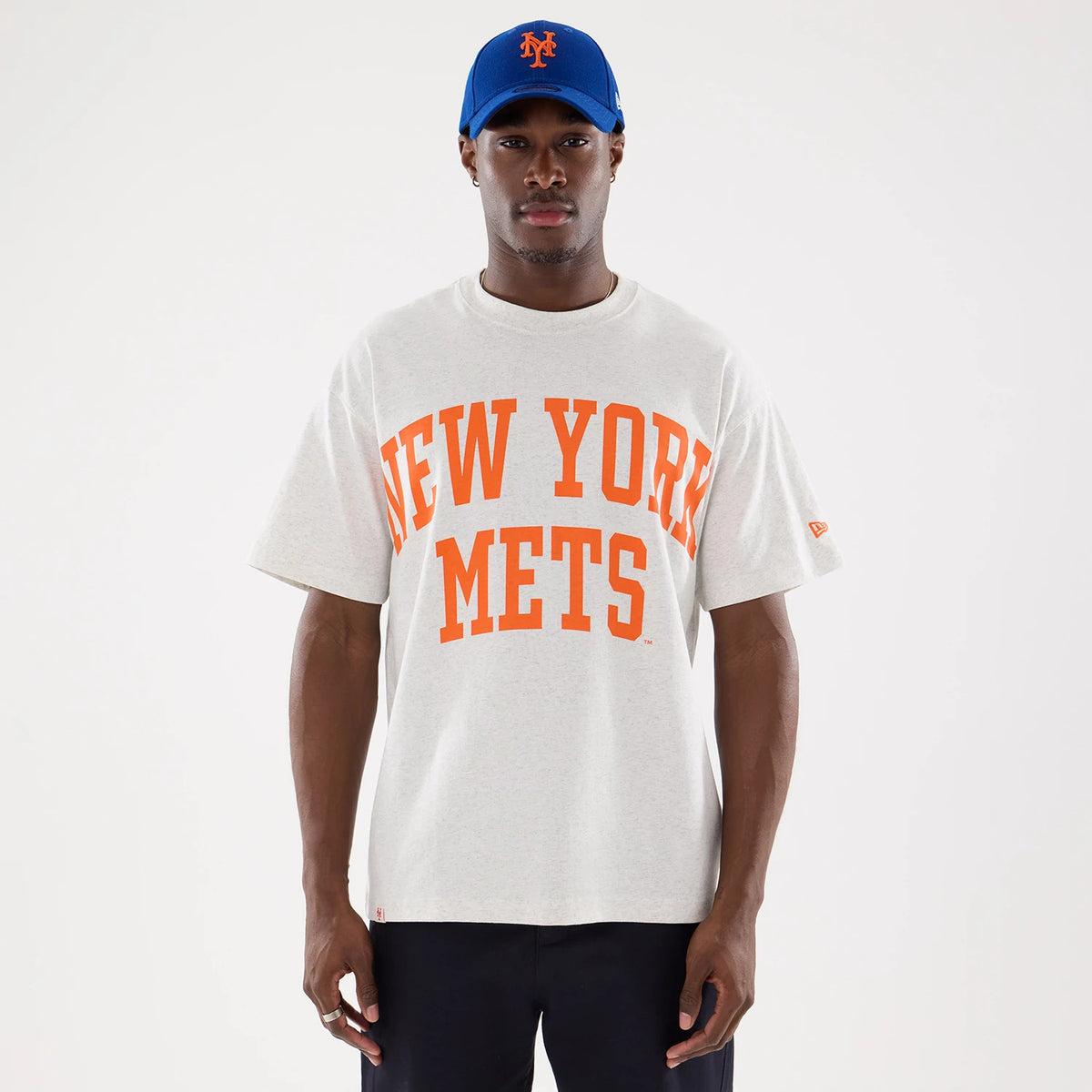 New York Mets MLB Comfort Grey T-Shirt, on-model view showing the light grey shirt with bold orange "New York Mets" text across the chest, a small embroidered New Era logo on the left sleeve, paired with a blue cap featuring a raised Mets logo.