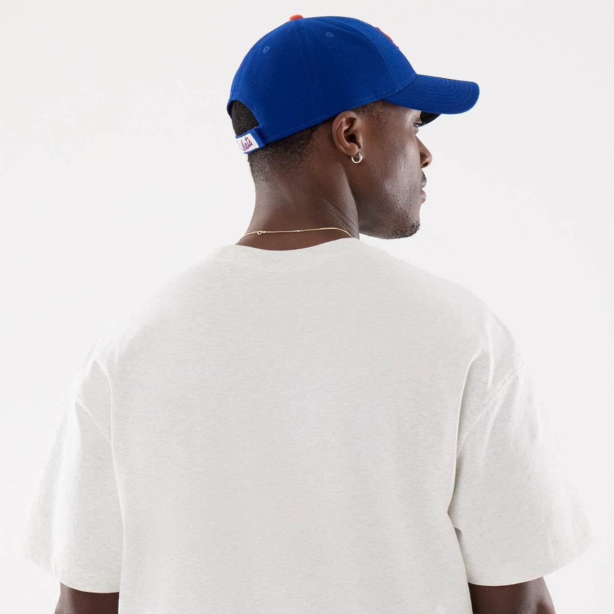 New Era New York Mets MLB Comfort Grey T-Shirt, on-model rear view featuring a plain grey tee extending across the shoulders and back, a blue cap with a white debossed logo above the adjustable strap at the back and a row of eyelets on top.