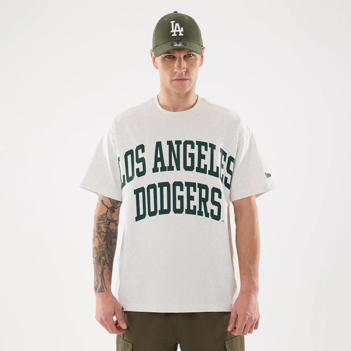 LA Dodgers MLB Comfort Grey T-Shirt, front view showing bold dark green "Los Angeles Dodgers" text across the chest, worn by a model with a green cap featuring a raised white LA logo on the front.