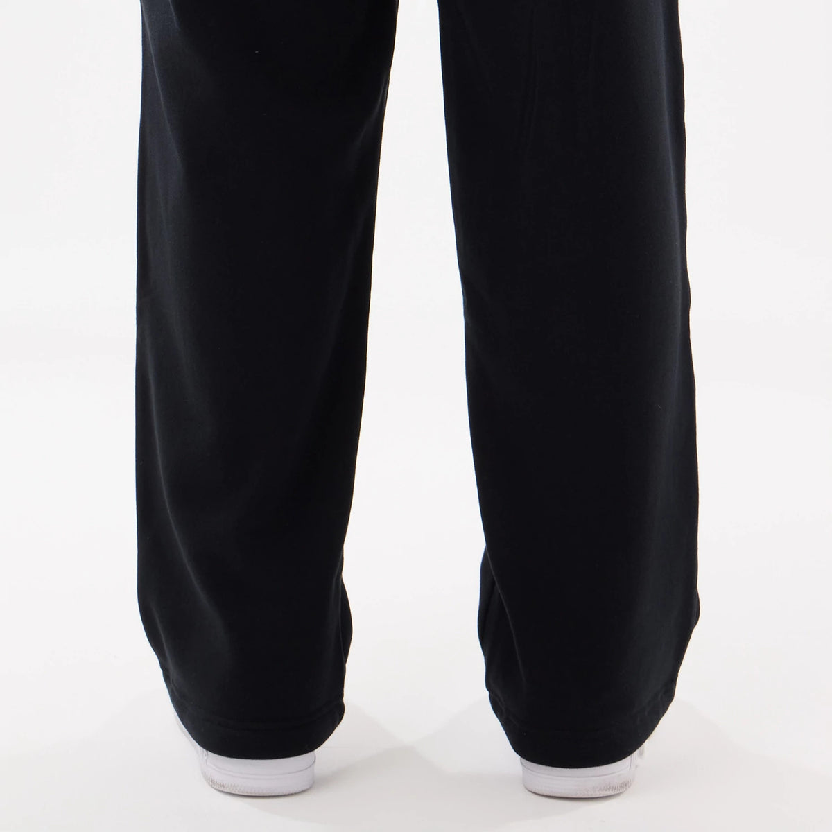 New Era Black Straight Legged Joggers, rear view showing relaxed fit leg openings draping over white trainers, with simple seam lines running vertically down the lower part of the black fabric.