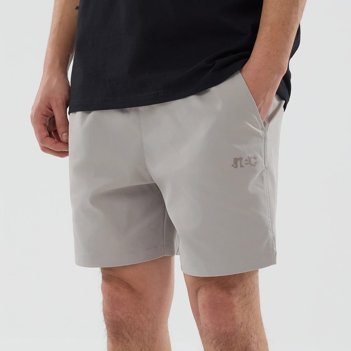 New Era NEC Logo Beige Oversized Shorts, front view showing the light beige fabric with a raised NEC logo on the left thigh, a relaxed fit falling above the knee and hands placed in the shorts' side pockets.