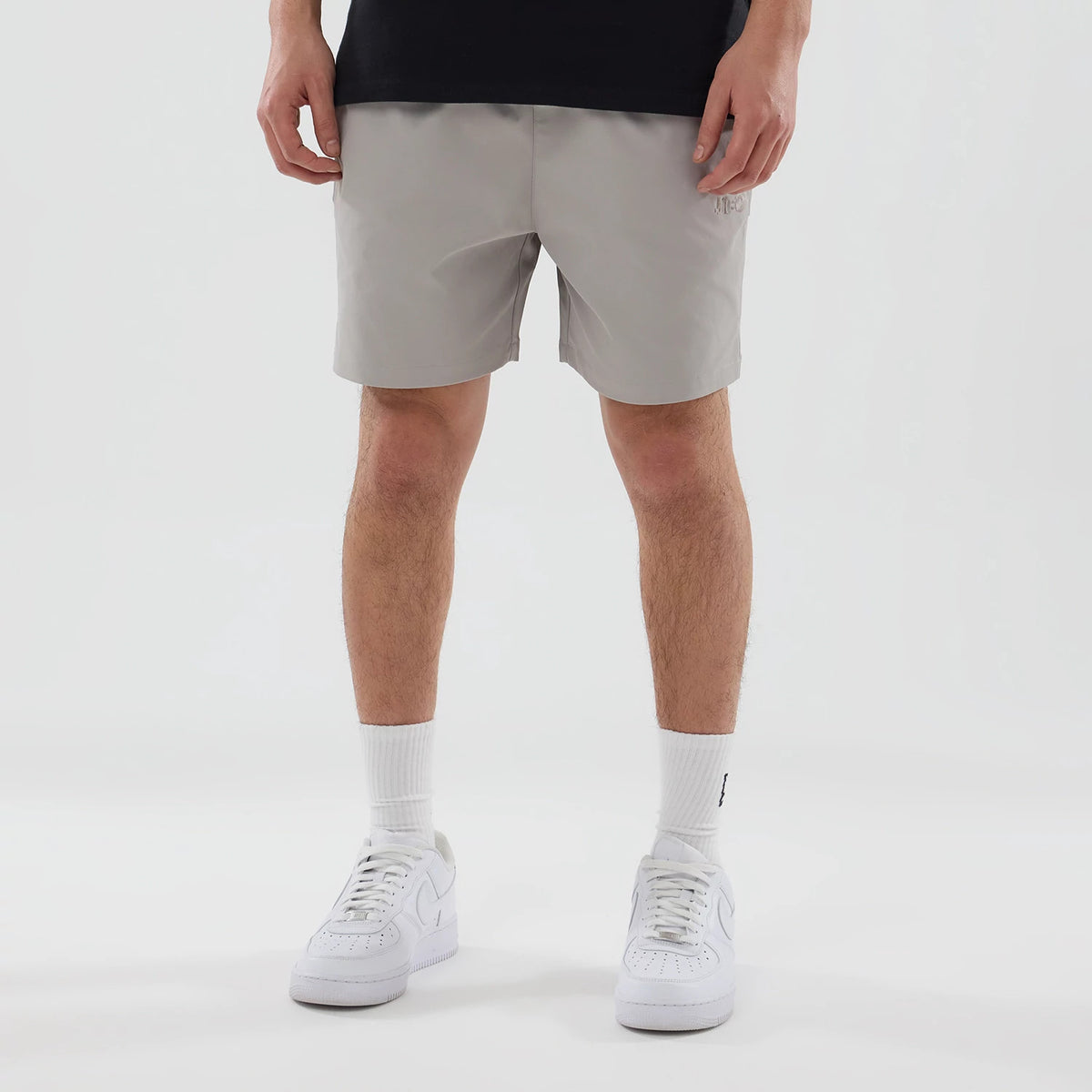 New Era NEC Logo Beige Oversized Shorts, on-model view showing the relaxed fit beige shorts with a raised NEC logo on the left leg, hem falling above the knee, paired with white trainers and socks.