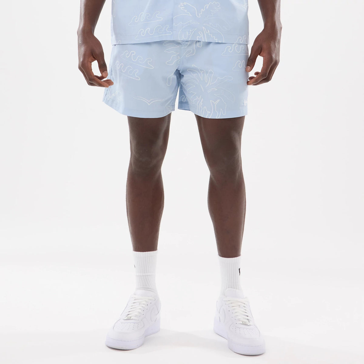 New Era Resort Blue Oversized Shorts, front view showing light blue shorts with white wave and palm tree patterns, reaching mid-thigh, worn with a matching blue shirt, and white sneakers completing the outfit.