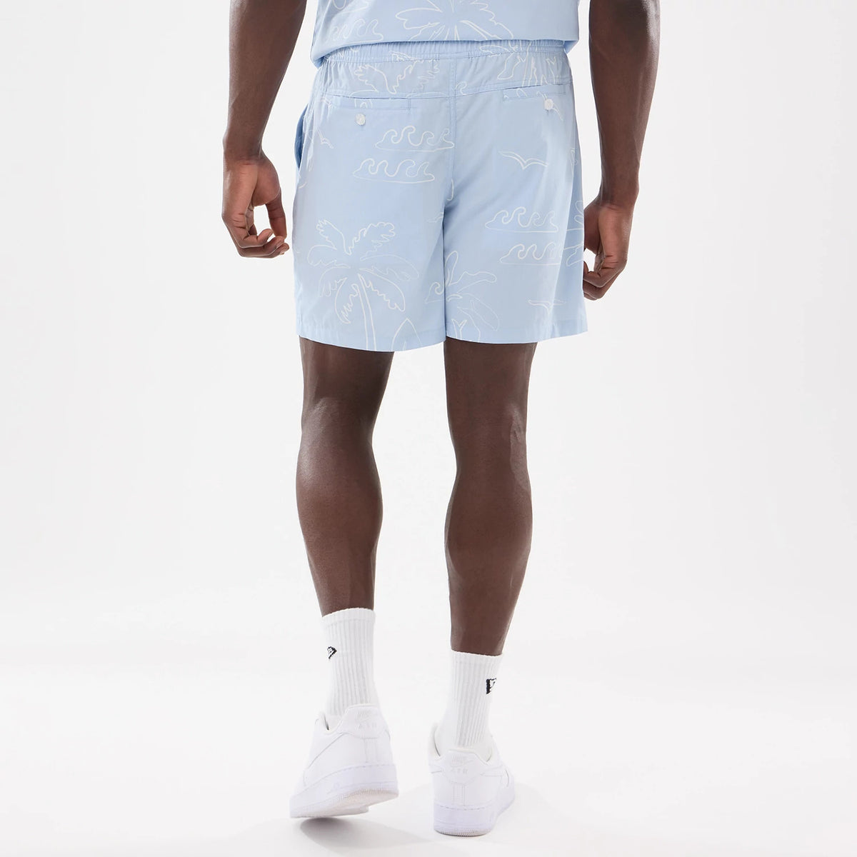 New Era Resort Blue Oversized Shorts, rear view showing a light blue fabric with white palm tree and wave patterns, an elastic waistband, and side pockets, worn by a person standing.