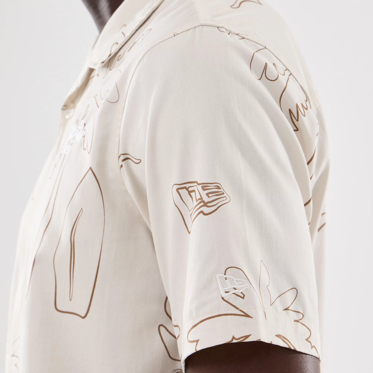 New Era Resort Natural Short Sleeve Shirt, side view showing intricate brown line art patterns on a natural background with a prominent New Era flag logo embroidered in white on the left sleeve.