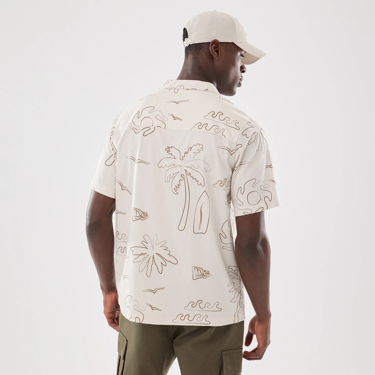 New Era Resort Natural Short Sleeve Shirt, back view on a model wearing a light cap, features a brown line-drawn pattern of palm trees, surfboards, and waves, covering the ivory fabric and extending across the back.