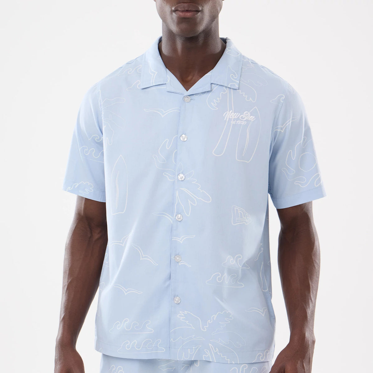 New Era Resort Blue Short Sleeve Shirt, front view on a model showing a white embroidered New Era logo on the left chest, buttons down the centre, and a pattern of abstract summer-themed outlines.