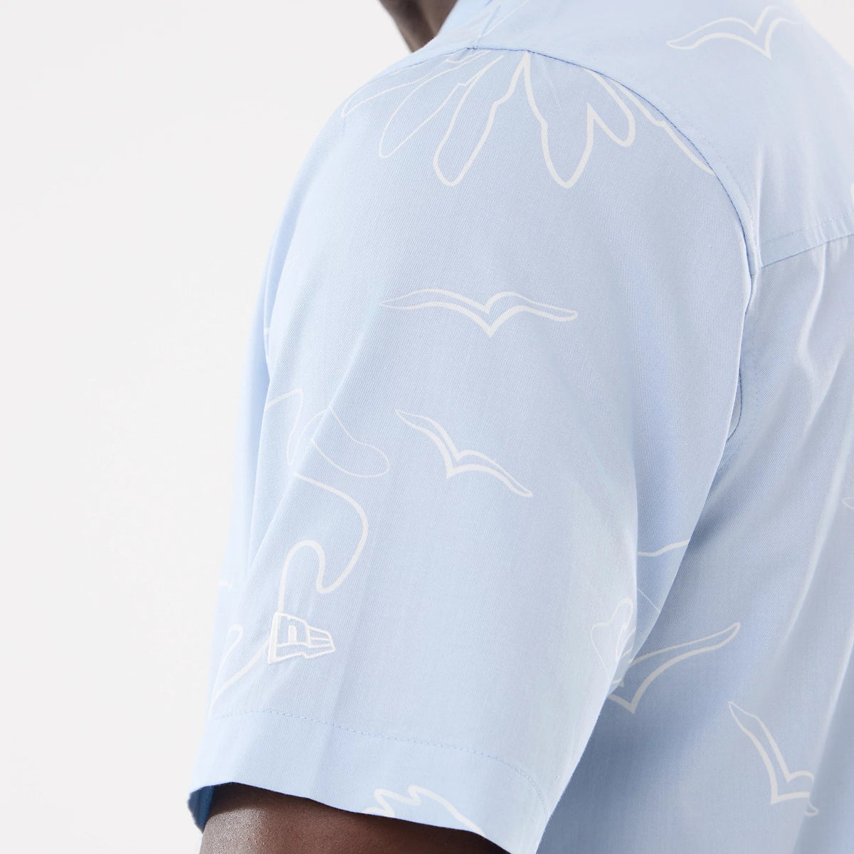 New Era Resort Blue Short Sleeve Shirt, side view on a model showing white seagull and wave patterns across the light blue fabric, with the New Era flag logo on the left sleeve near the hem.