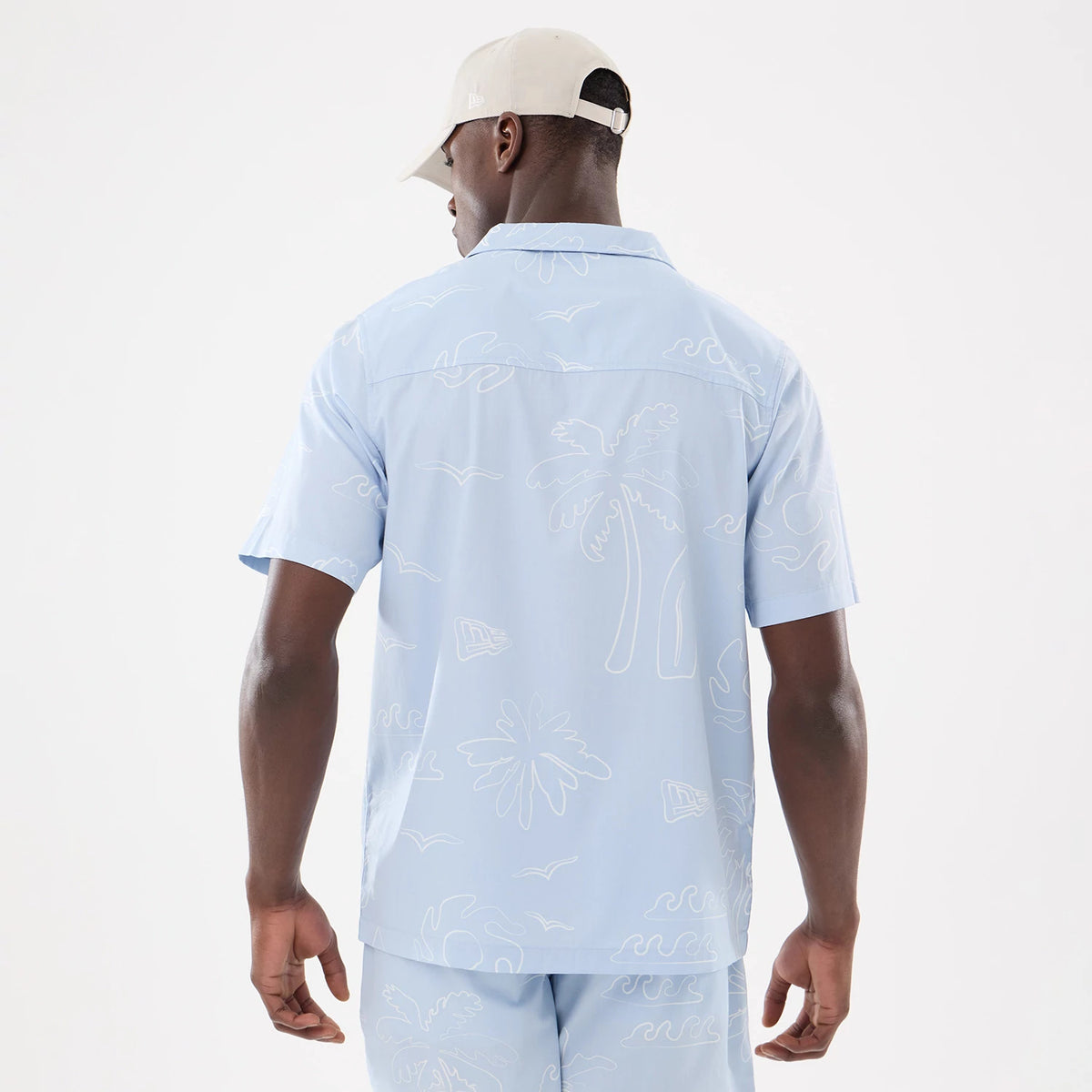 New Era Resort Blue Short Sleeve Shirt, back view on a model wearing matching shorts and a cream cap, showing white palm tree and wave patterns across the fabric with a relaxed straight hem and short sleeves.
