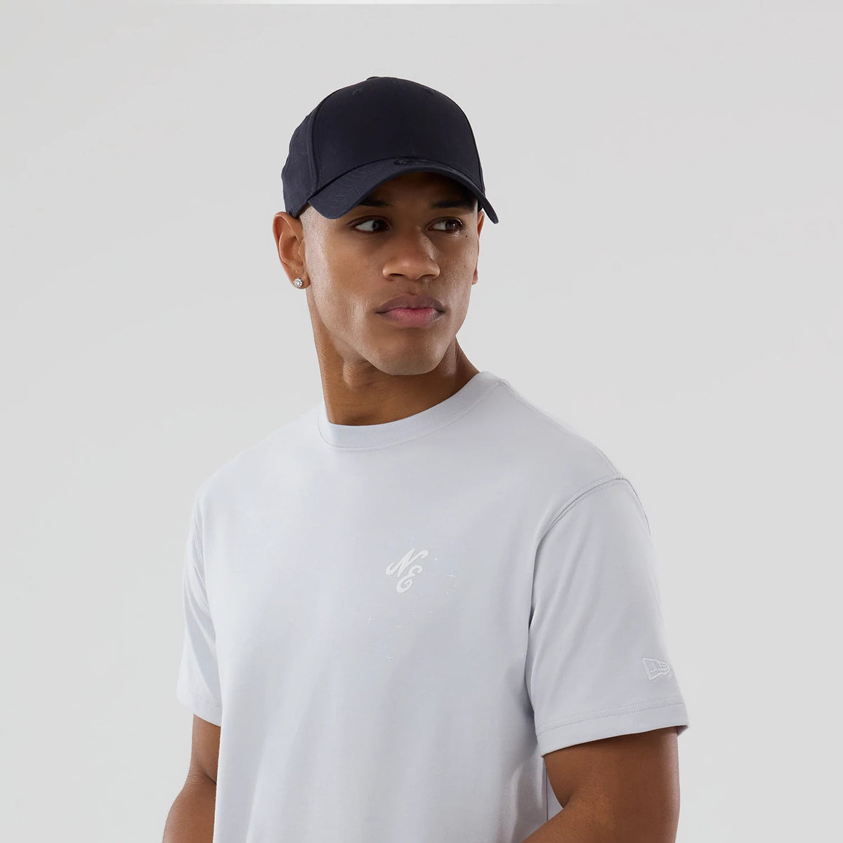 New Era Zodiac Sign Silver T-Shirt, on-model view showing a light grey T-shirt with a small white embroidered logo on the chest and another debossed New Era logo on the left sleeve, paired with a black cap.