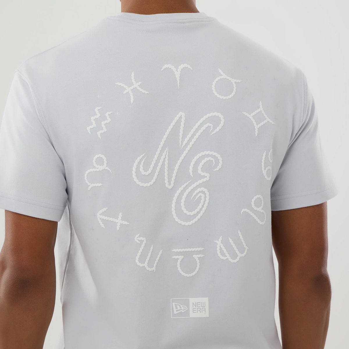 New Era Zodiac Sign Silver T-Shirt, rear view showing a white embroidered circle of zodiac symbols around a large "NE" on the upper back, with a debossed New Era logo near the bottom hem.