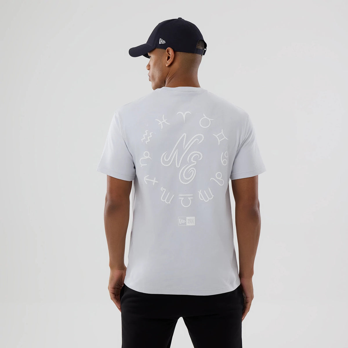 New Era Zodiac Sign Silver T-Shirt, rear view showing an embroidered pattern of zodiac symbols encircling stylised initials NE in silver, with a debossed New Era logo near the lower back hem, worn by a man with a black cap.