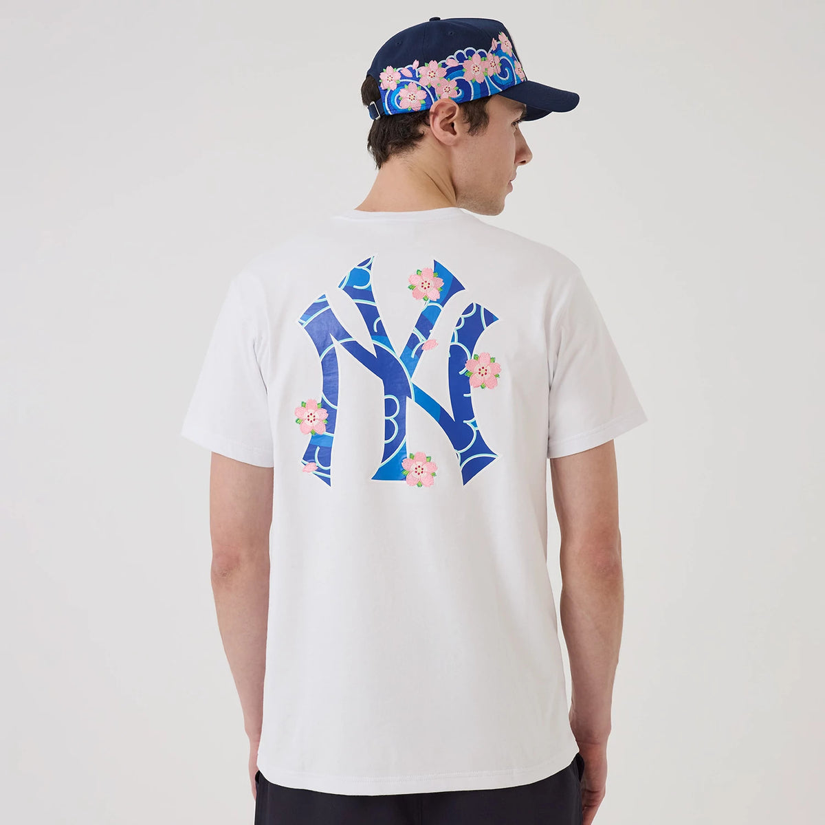 New York Yankees MLB Sakura White T-Shirt, rear view showing a large blue Yankees logo with pink floral accents on the back, worn by a model also in a navy cap featuring a matching floral pattern around the band.