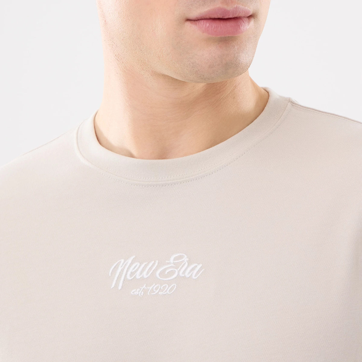 New Era Resort Natural T-Shirt, on-model view showing the upper section of a light cream t-shirt with a raised white New Era logo and "est. 1920" text centred on the chest below the round neckline.