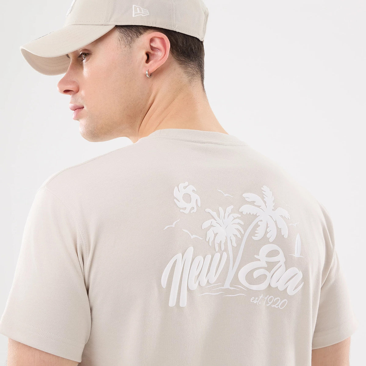 New Era Resort Natural T-Shirt, on-model rear view showing a light-coloured design with a raised logo featuring palm trees and birds, the model in a matching cap with a small embroidered logo on the side.