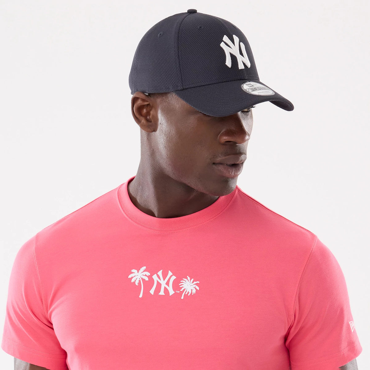 New York Yankees MLB Resort Pink T-Shirt, on-model view showing the bright pink fabric with a white palm tree and Yankees logo design on the chest, paired with a navy cap featuring a raised white Yankees logo on the front.