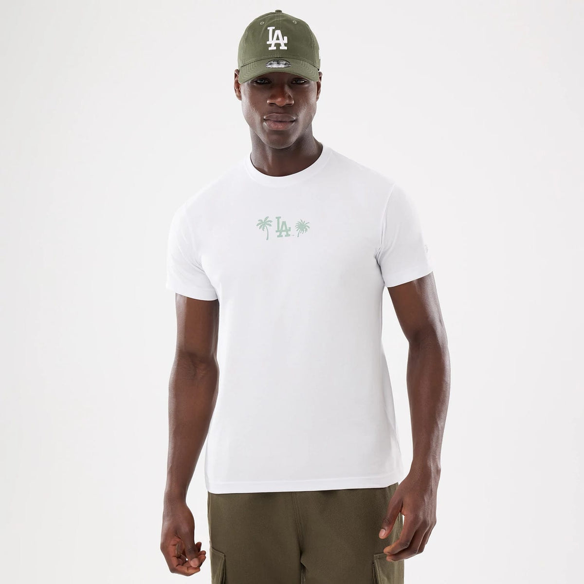 LA Dodgers MLB Resort White T-Shirt, front view showing a white T-shirt with a light green "LA" logo and palm tree graphics on the chest, worn by a model with a green cap featuring a raised "LA" logo.