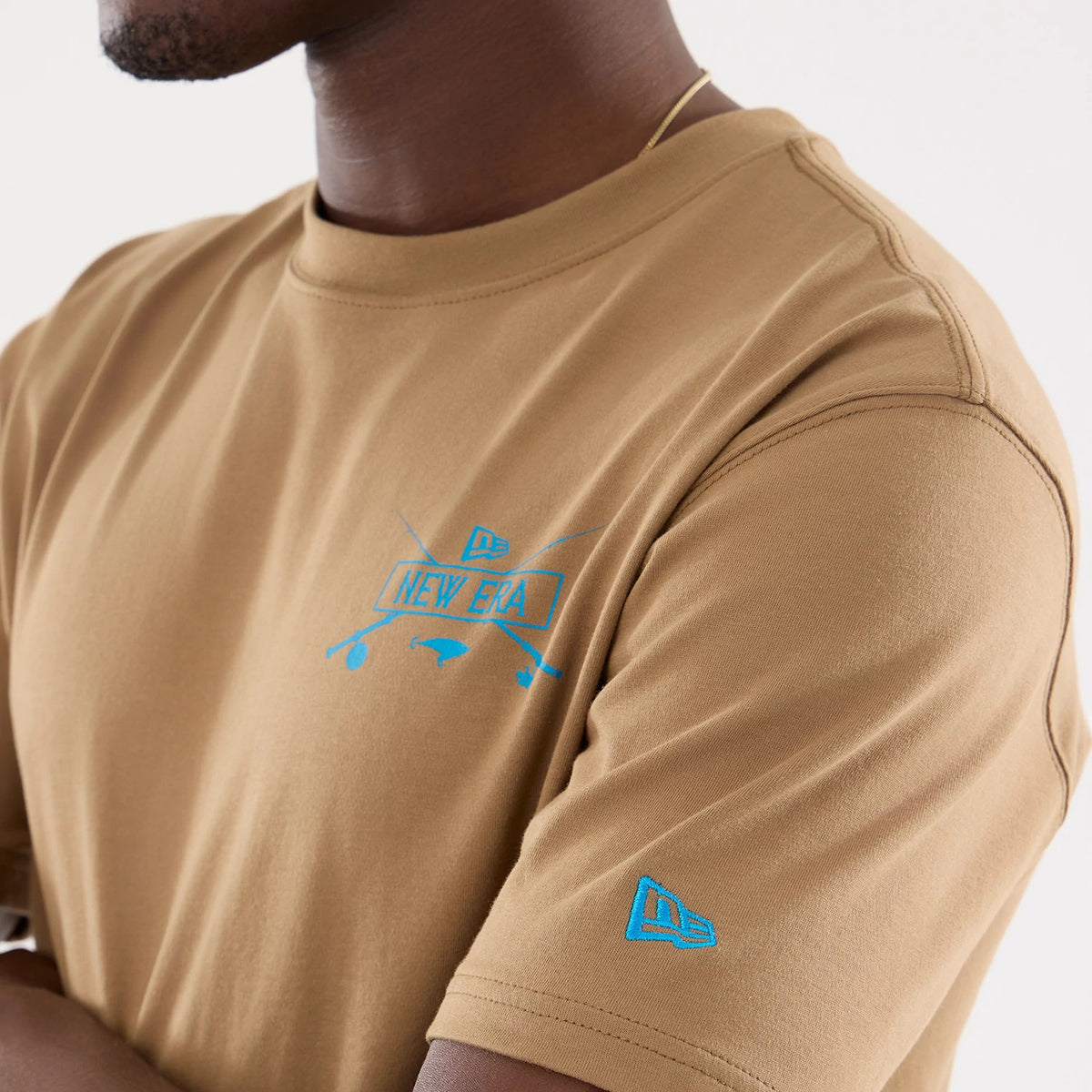 New Era Graphic Outdoor Fishing Beige T-Shirt, front view showing the short-sleeve design with a blue raised New Era fishing graphic on the chest and the raised logo on the left sleeve, all on a beige fabric.