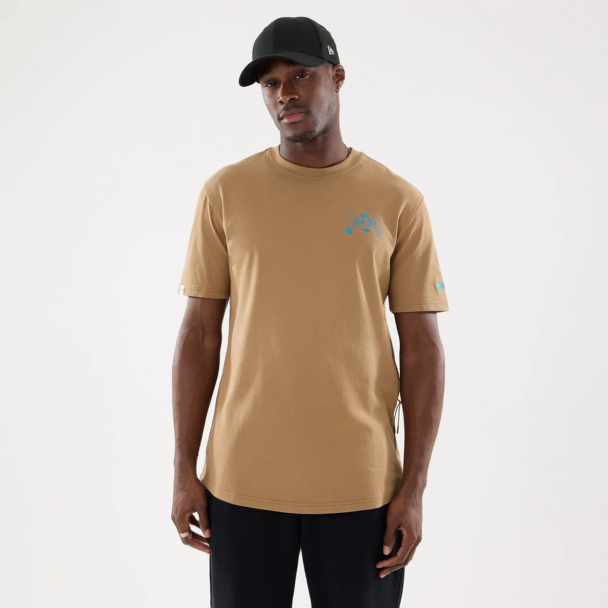 New Era Graphic Outdoor Fishing Beige T-Shirt, front view showing a beige short-sleeve design with a blue graphic logo on the chest and a small logo on the left sleeve, worn by a model wearing a black cap.