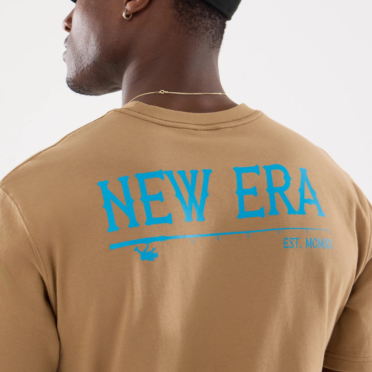 New Era Graphic Outdoor Fishing Beige T-Shirt, rear view showing a large blue New Era graphic across the upper back, with a fishing rod element below and the text "EST. MCMXX" to the right against the beige fabric.