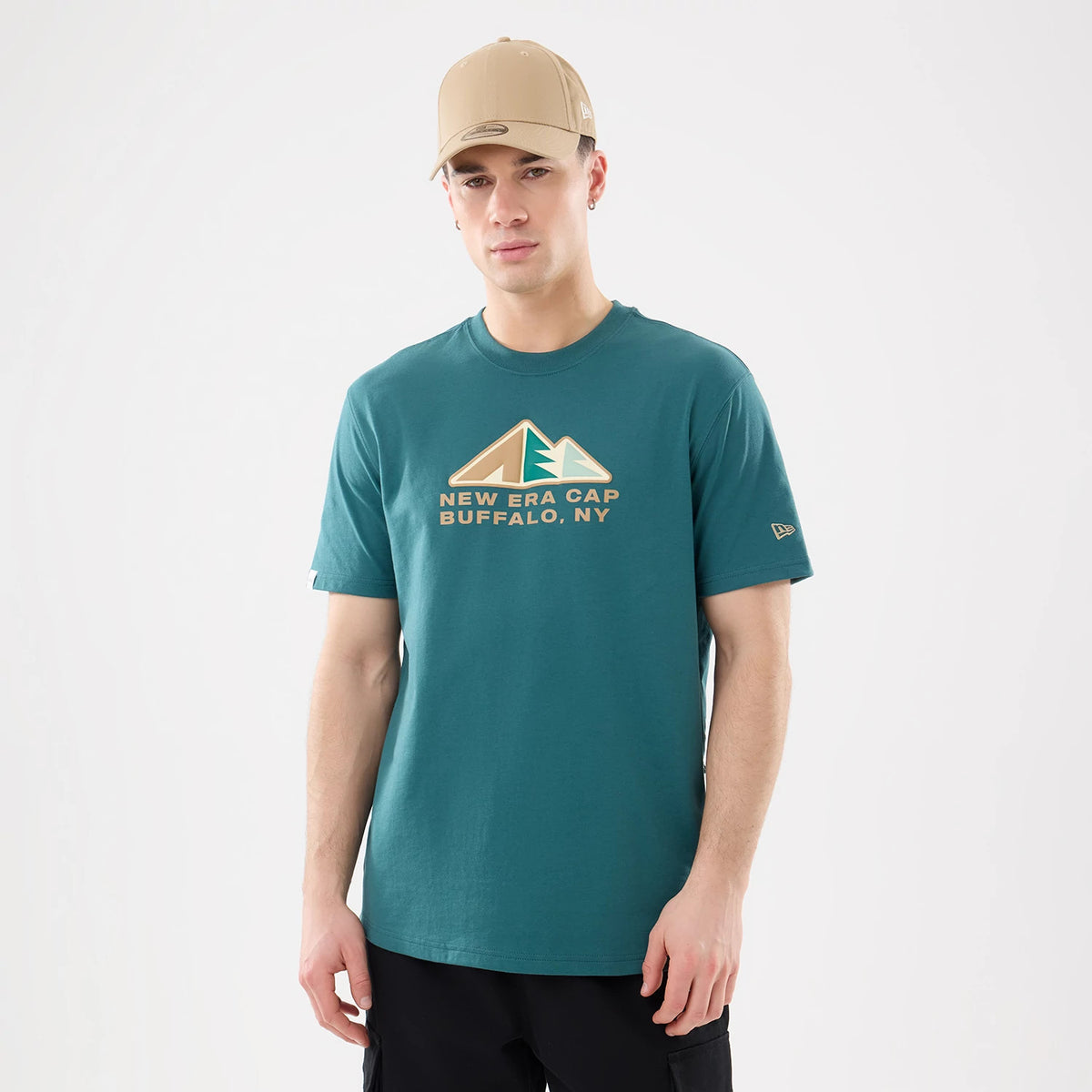 New Era Outdoor Mountain Dark Green T-Shirt, on-model view showing a dark green shirt with a large mountain graphic and text "NEW ERA CAP BUFFALO, NY" across the chest, a debossed New Era logo on the left sleeve, worn with a tan cap.