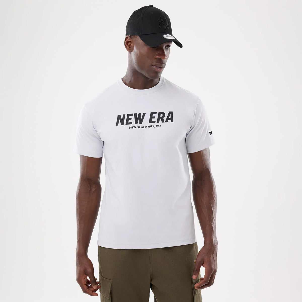 New Era Basic White T-Shirt, on-model front view showing a white T-shirt with a bold black printed "NEW ERA" logo and "BUFFALO, NEW YORK, USA" text across the chest, worn by a person in a black cap with a raised logo.