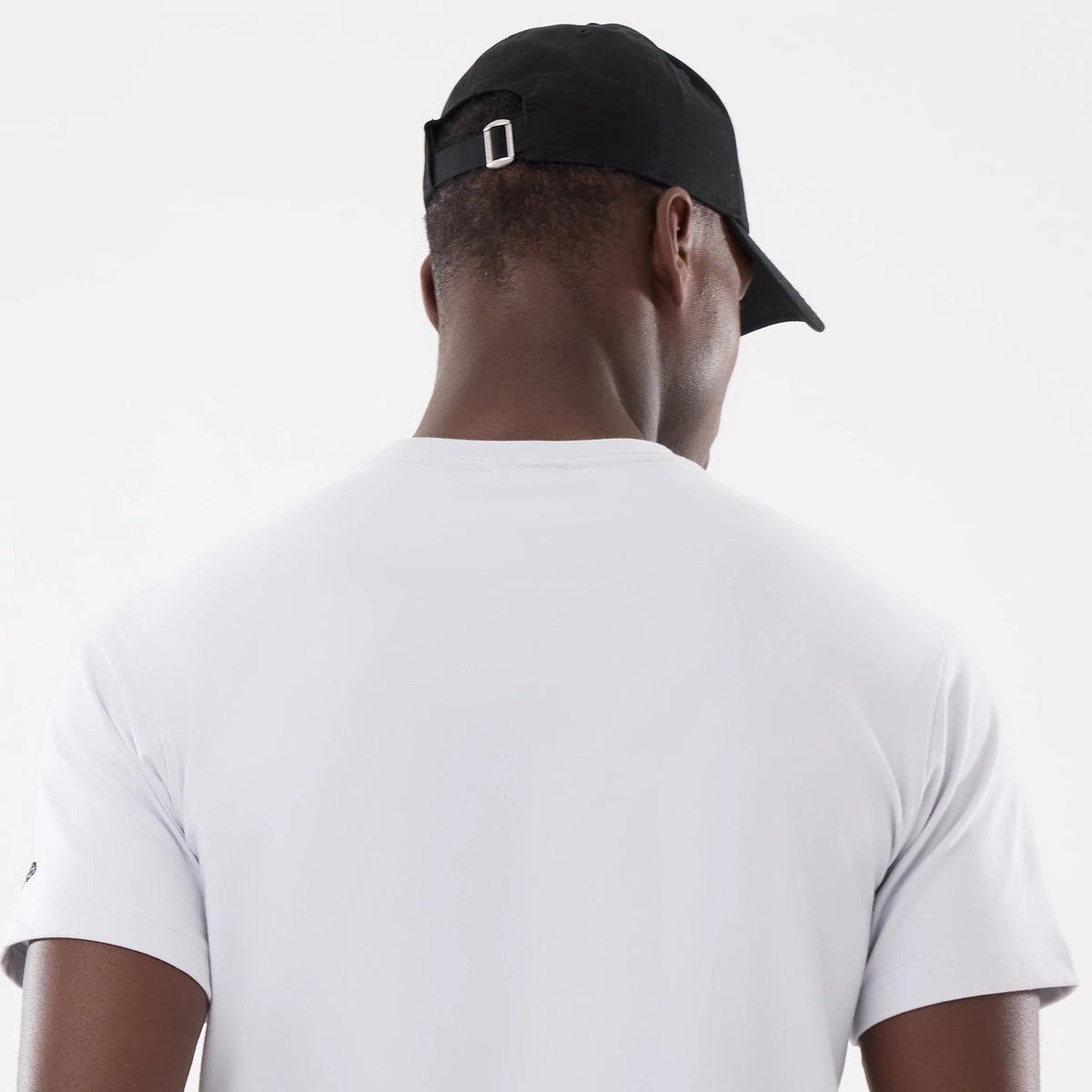 New Era Basic White T-Shirt, rear view showing the plain white fabric extending over the shoulders, sleeves slightly folded over the arm, and a black cap on-model with a silver buckle on the back strap.