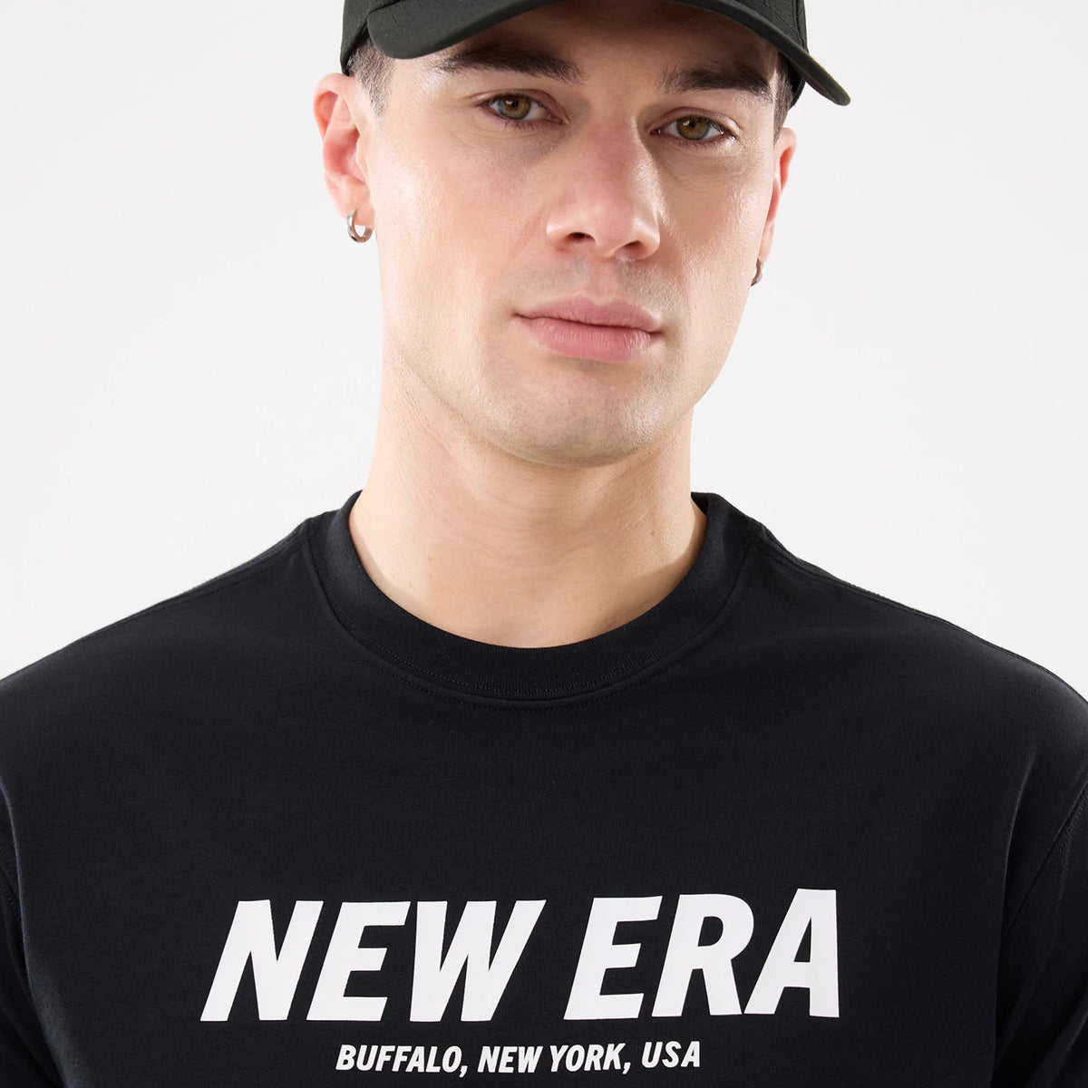 New Era Basic Black T-Shirt, on-model view showing the front with bold white "NEW ERA" text across the chest, accompanied by "BUFFALO, NEW YORK, USA" below, worn by a person also in a black cap.