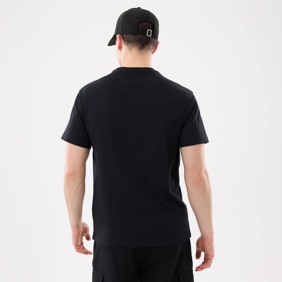 New Era Basic Black T-Shirt, rear view showing the plain black shirt worn on-model, highlighting the straight hem and short sleeves, paired with a black cap featuring an embossed New Era logo on the back.