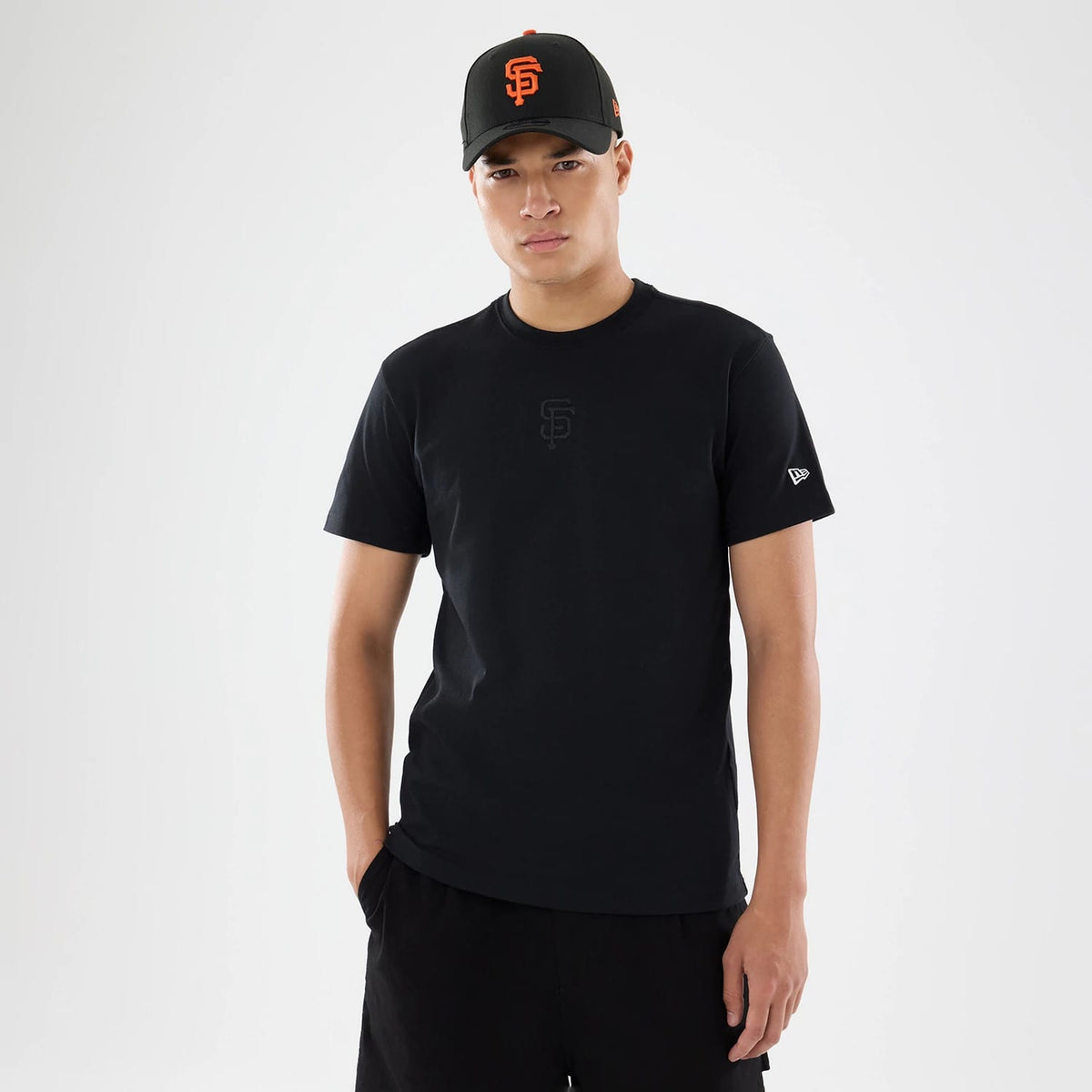 San Francisco Giants MLB Cartoon Black T-Shirt, on-model view showing a black T-shirt with an embossed black emblem on the chest and a raised New Era logo on the sleeve, paired with a black cap featuring an embossed orange emblem on the front.