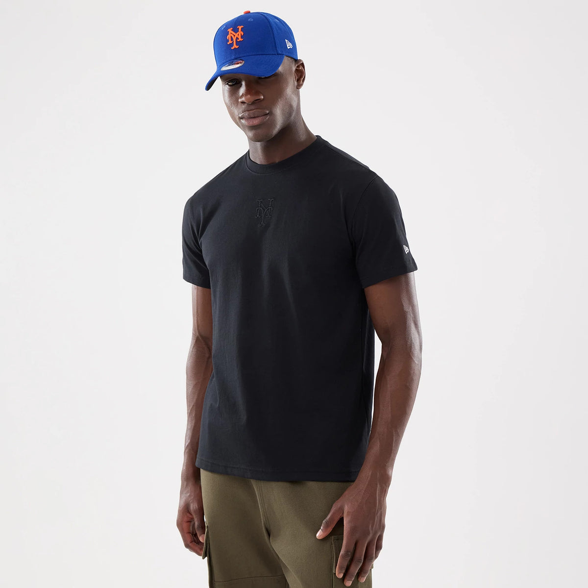 New York Mets MLB Cartoon Black T-Shirt, on-model view showing the black shirt with an embossed logo on the chest, worn with a blue cap that has a raised orange logo, and small debossed logo on the sleeve.