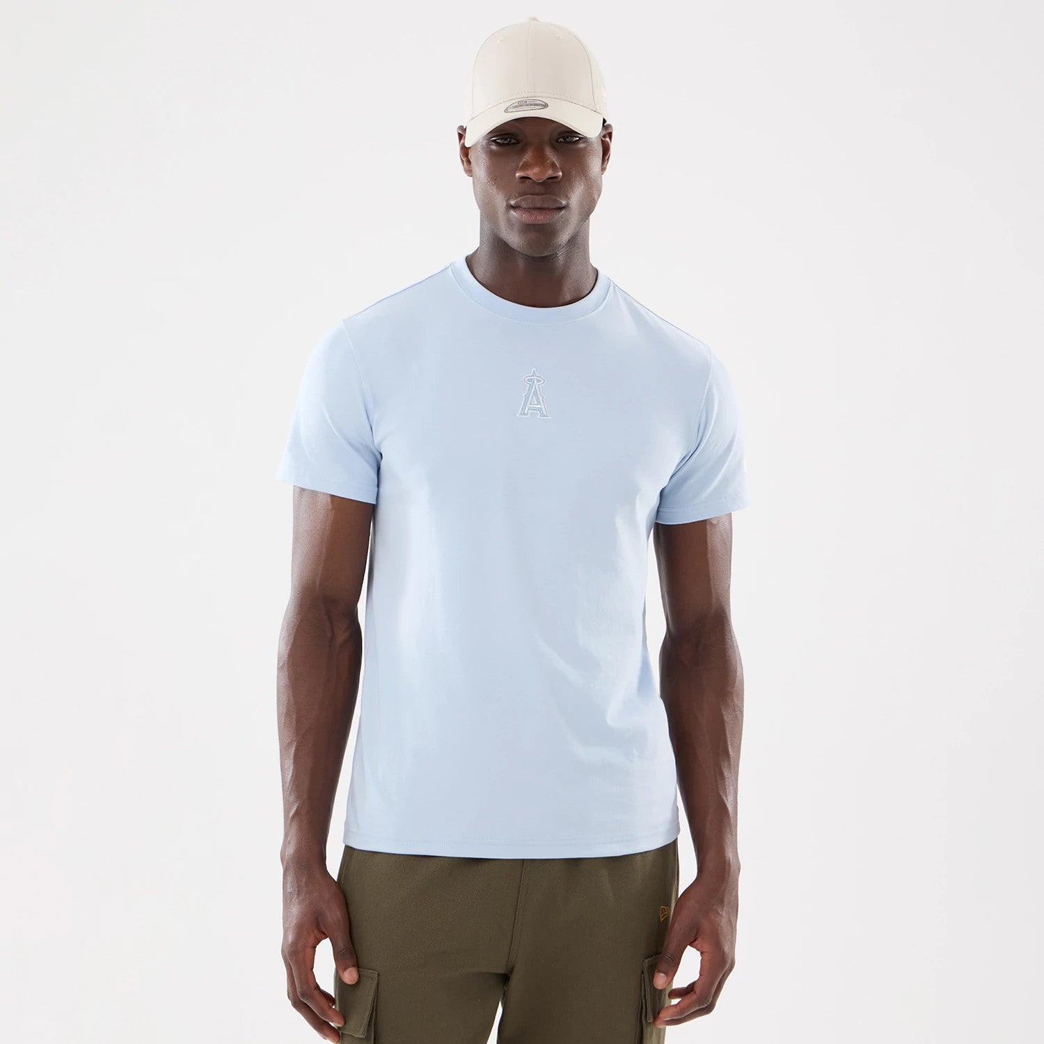 New Era LA Angels MLB Cartoon Blue T-Shirt, front view showing a light blue T-shirt with a raised LA Angels logo centred below the neckline, worn by a model also wearing a beige cap and olive green trousers.