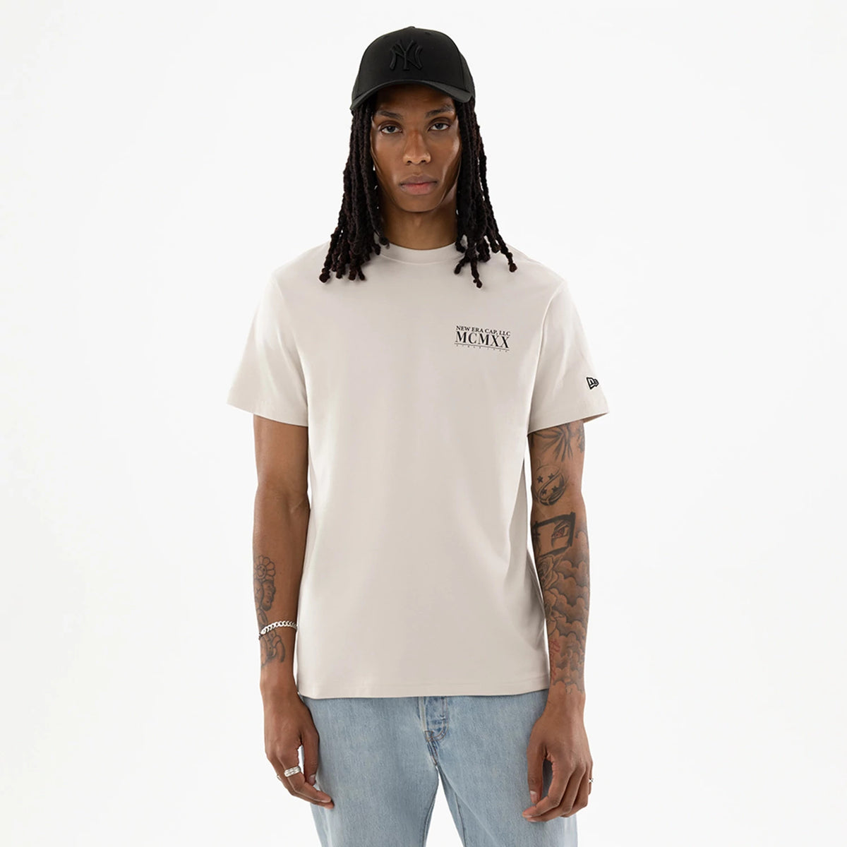 New Era Heritage Cream T-Shirt, front view showing a cream T-shirt with black "MCMLXX" text on the left chest, black raised New Era logo on a black cap worn on the model, and a small raised New Era logo on the left sleeve.