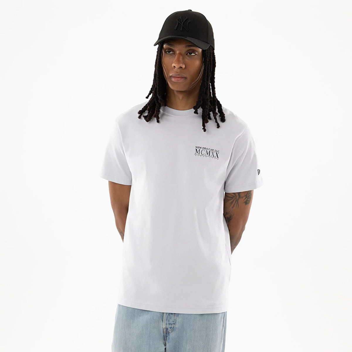 New Era Heritage White T-Shirt, on-model view showing the short-sleeved white t-shirt with "NEW ERA CAP CO MCMXX" printed on the left chest, a small logo on the sleeve, paired with a black cap featuring a raised logo.