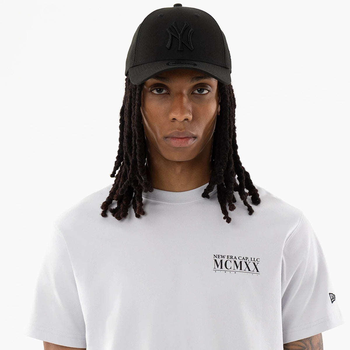 New Era Heritage White T-Shirt, front view of the white T-shirt featuring a black-printed logo with text on the left chest, worn by a model in a black cap with a raised logo and eyelets on the front panels.