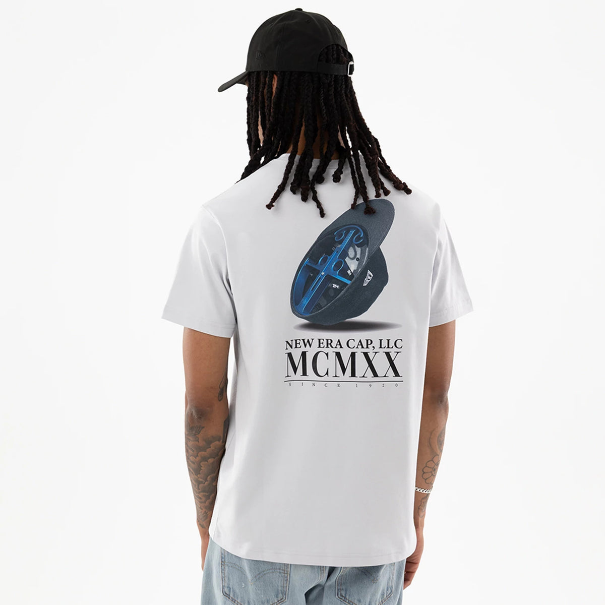 New Era Heritage White T-Shirt, back view showing a graphic of a black cap with a blue design on the crown, text reading "NEW ERA CAP, LLC MCMXX" below, worn by a model in blue jeans.