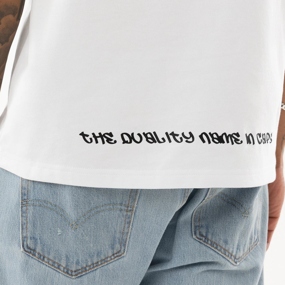 New Era I Am Original White T-Shirt, on-model view showing the lower back of the white T-shirt with the black text “the quality name in caps” printed along the hem above light blue denim jeans.