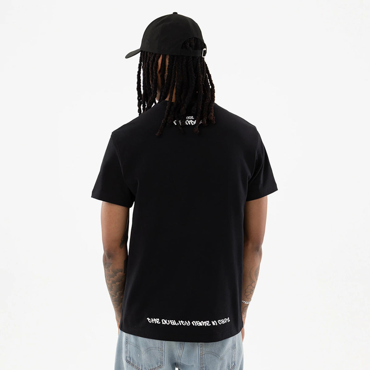 New Era I Am Original Black T-Shirt, rear view showing the black fabric with white slogan text near the hem, a small white embroidered detail beneath the collar, and a man wearing a black cap with a debossed logo.