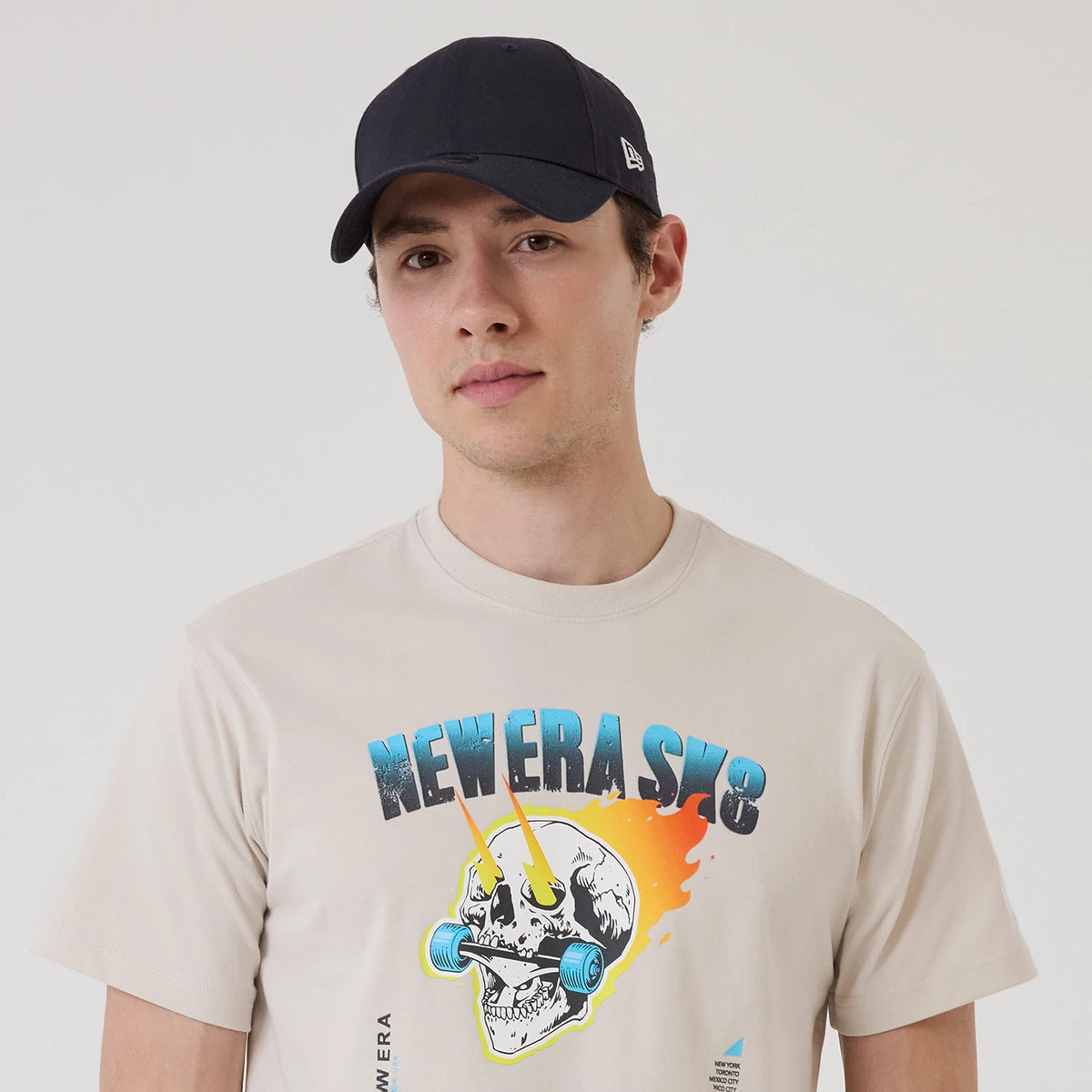 New Era SK8 Graphic Cream T-Shirt, on-model view showing a vivid skull graphic with skateboard wheels under a New Era SK8 logo in blue and black, worn by a person in a black cap with an embroidered logo on the side.