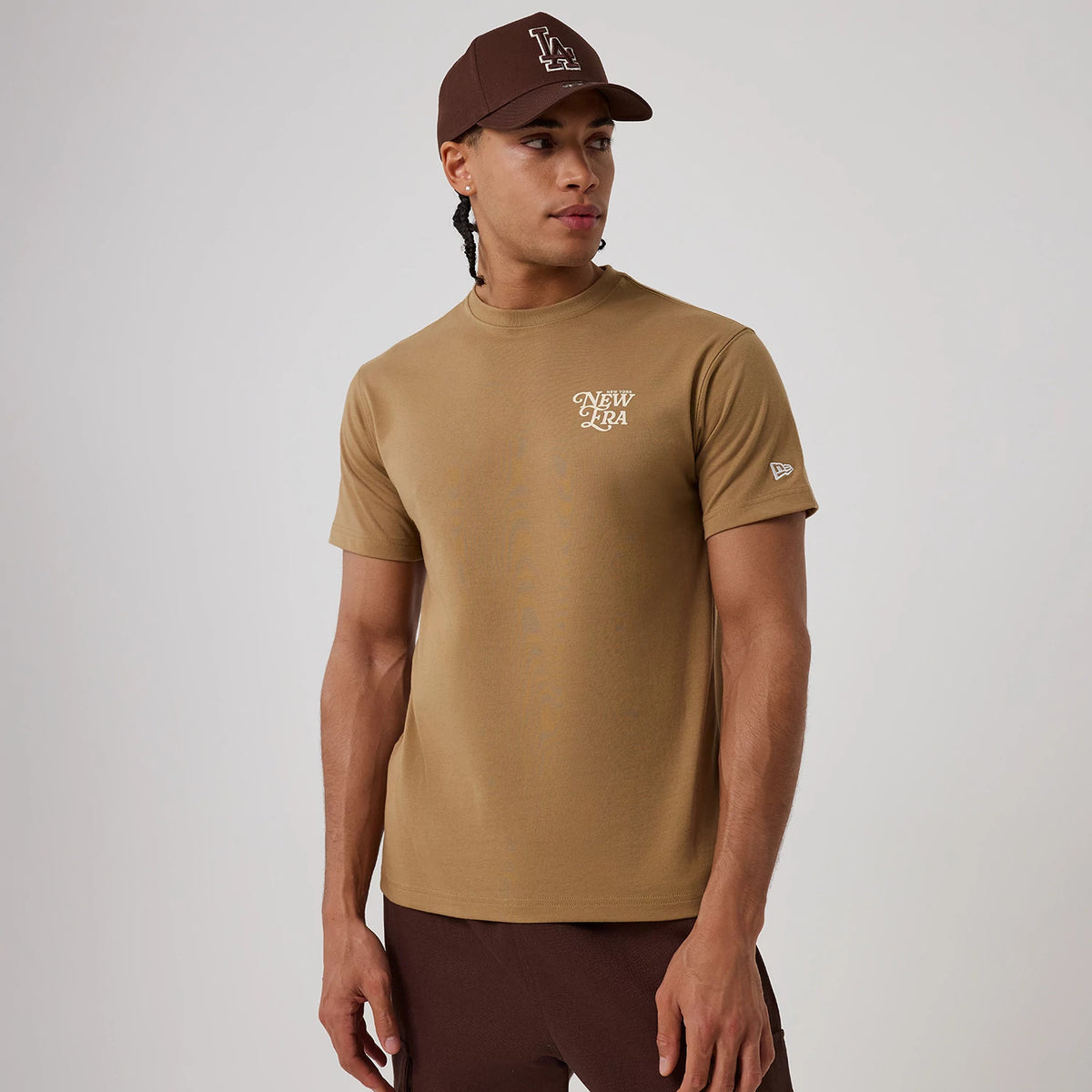 New Era Script Graphic Beige T-Shirt, on-model view showing a beige short-sleeve design with the white script New Era logo on the left chest, complemented by a brown cap featuring a white LA logo and a debossed New Era logo on the sleeve.