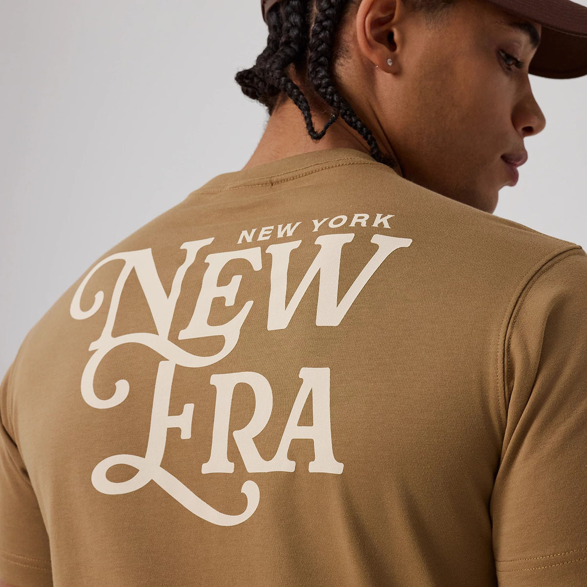 New Era Script Graphic Beige T-Shirt, on-model view featuring the oversized white New Era script graphic printed across the upper back on a beige background, worn by a person with braided hair wearing a brown cap.