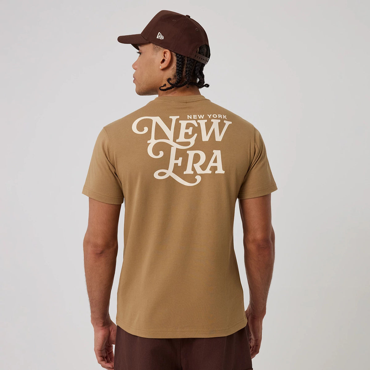 New Era Script Graphic Beige T-Shirt, back view showing a large white print with stylized lettering centred on the beige fabric, worn by a person also wearing a brown cap with a raised New Era logo on the side.
