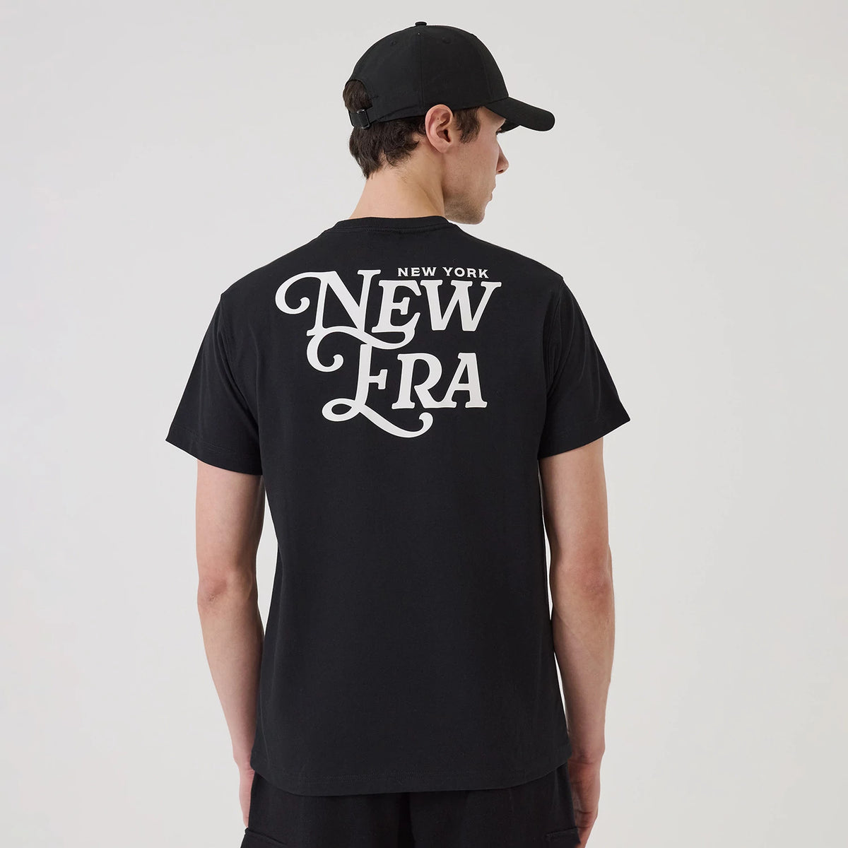 New Era Script Graphic Black T-Shirt, rear view featuring a large white script logo printed across the upper back, worn by a person also in a black cap, showcasing the casual fit of the short sleeves and straight hem.